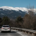 A white car driving on a curvy mountain road with snow-capped peaks in the background.